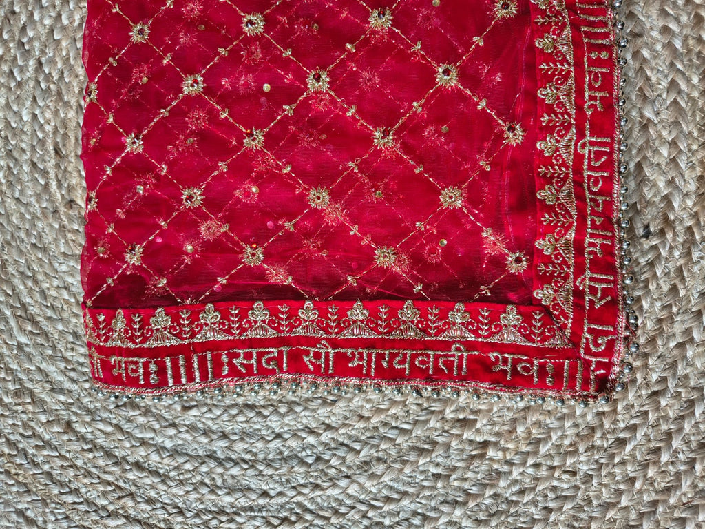 Corner detail of red net dupatta on jute showing Sada Saubhagyavati Bhava gold Sanskrit border and silver ghungroo bead lace trim on edges