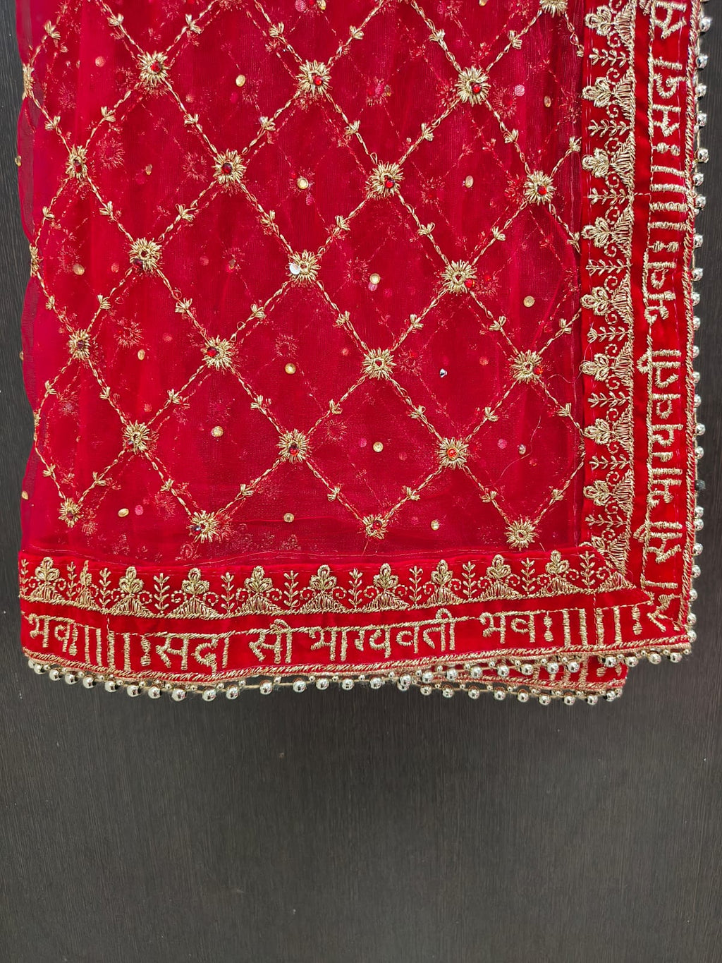 Close-up of red bridal dupatta border showing Sada Saubhagyavati Bhava gold Sanskrit embroidery on velvet strip with silver ghungroo bead lace trim