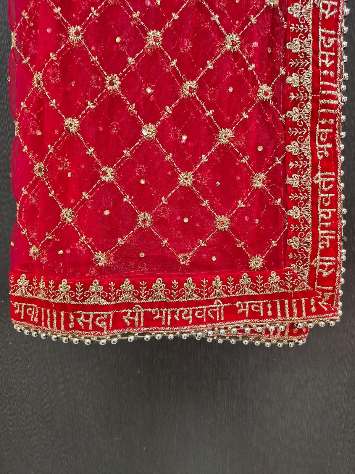 Close-up of red bridal dupatta border showing Sada Saubhagyavati Bhava gold Sanskrit embroidery on velvet strip with silver ghungroo bead lace trim